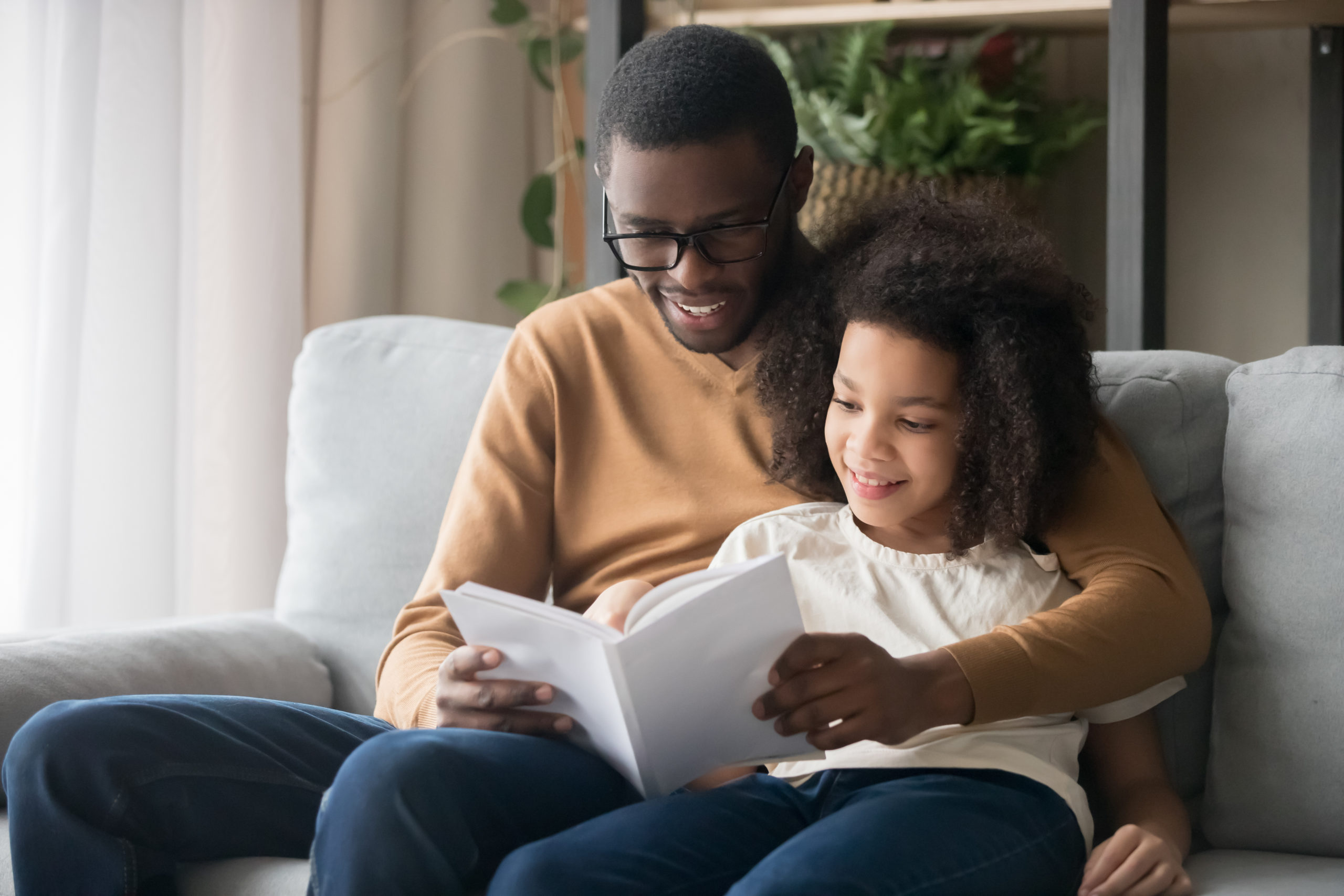 Happy family black father or baby sitter and kid daughter reading story book sitting on sofa together, african american child girl enjoying time with daddy listening to fairy tale from dad at home