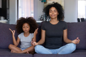 Black mother and little girl sitting in lotus pose on couch together, mum teaching child to meditate