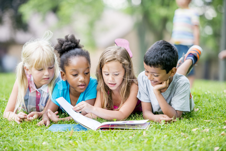 Three young girls and a boy are outdoors during summer. They are laying in the grass and reading a book together.